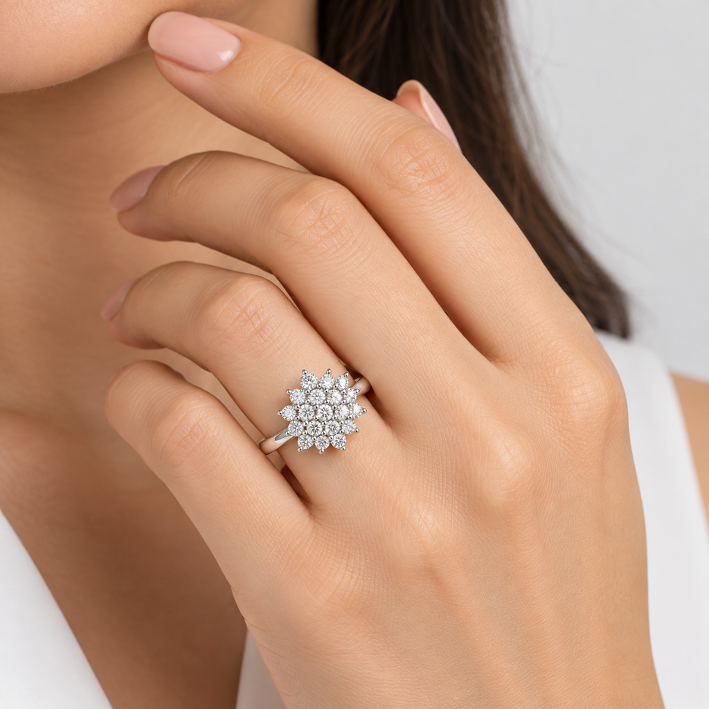 A close-up of a woman's hand wearing a silver-toned ring with a cluster of clear gemstones in a floral or snowflake-like design, against a soft light gray background.