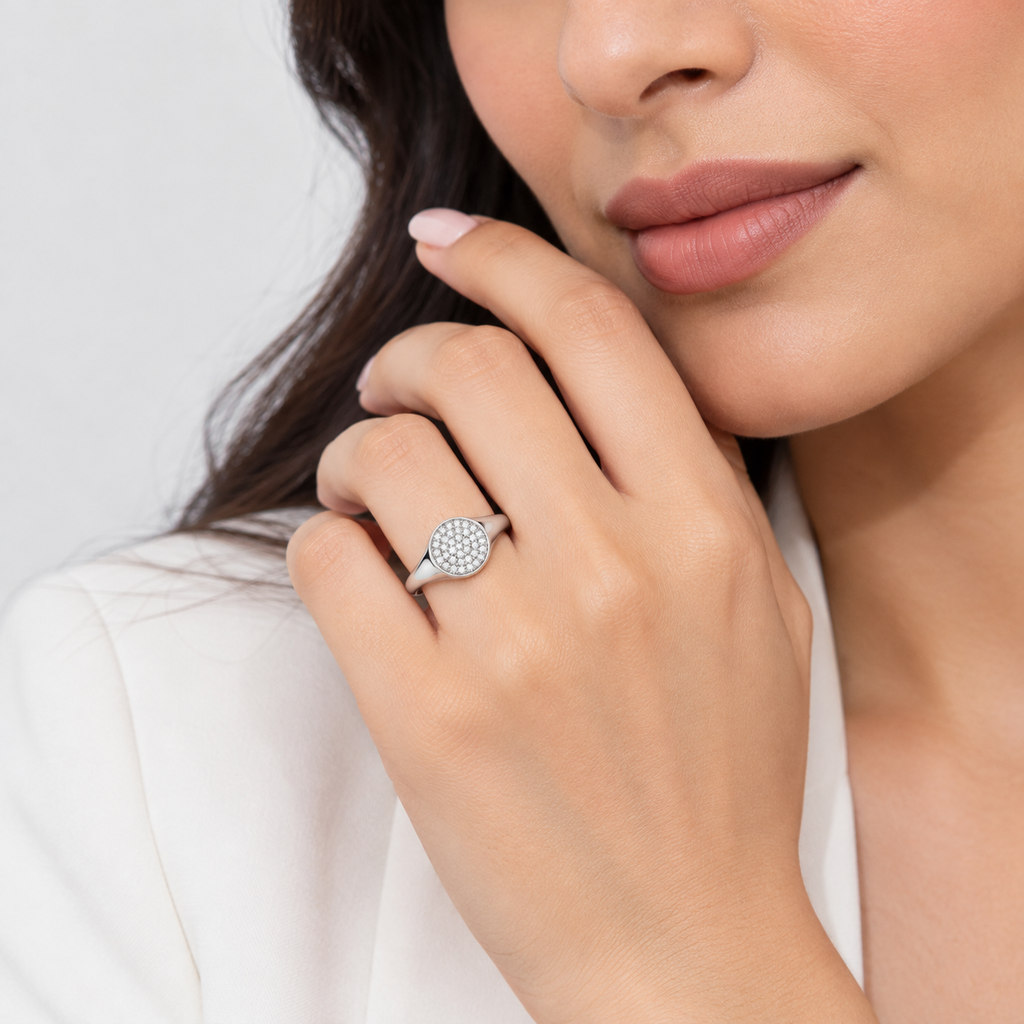 A close-up lifestyle shot of a woman wearing a silver ring with a round pavé diamond setting on her finger, dressed in a white blazer against a light gray background.