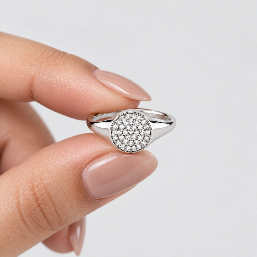 A silver signet ring with a round pavé-set face, held between fingers against a clean, light gray background.