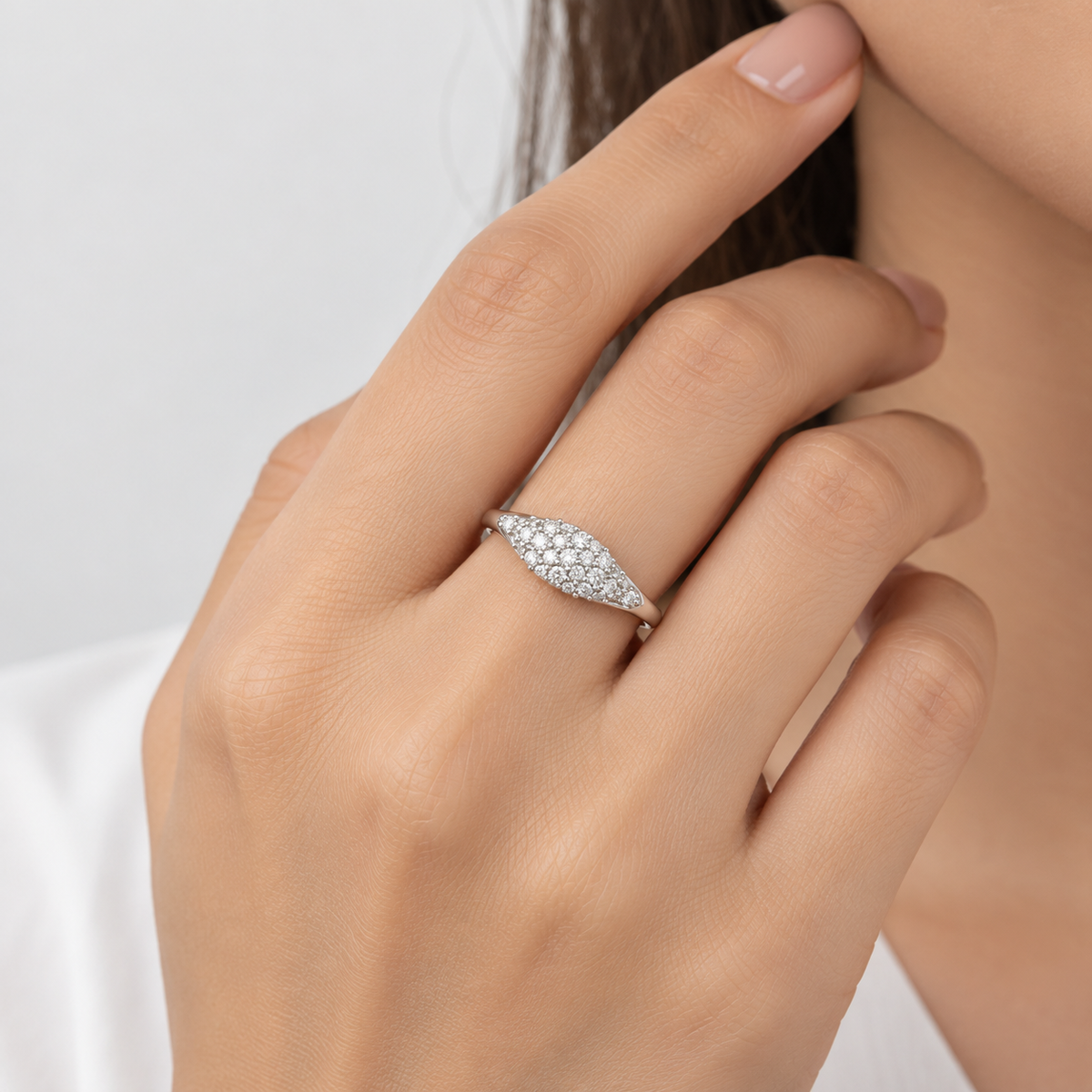 A close-up of a woman's hand wearing a silver ring with pavé-set clear stones, set against a soft, light gray background.