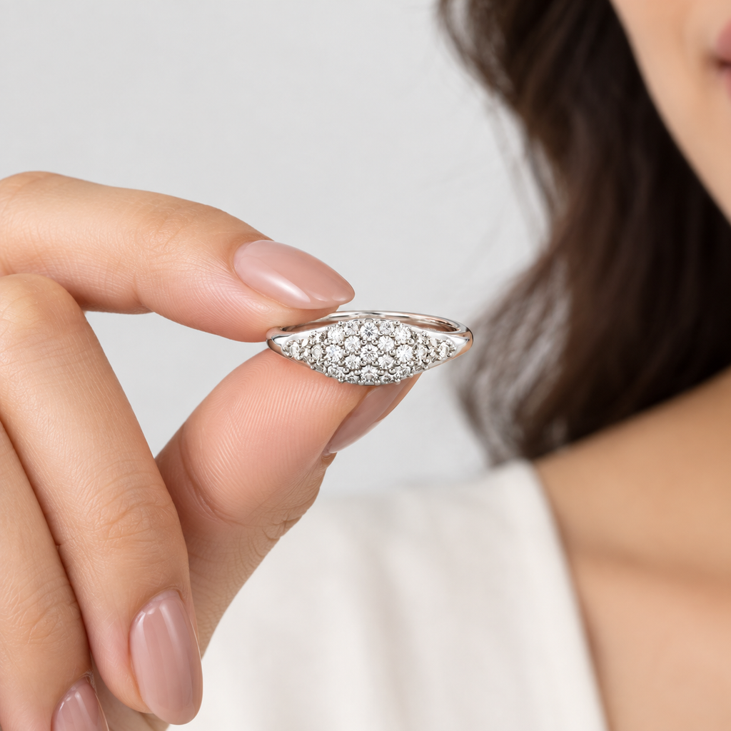 A close-up of a hand with manicured nails holding a silver ring adorned with pavé-set clear stones against a soft light gray background.