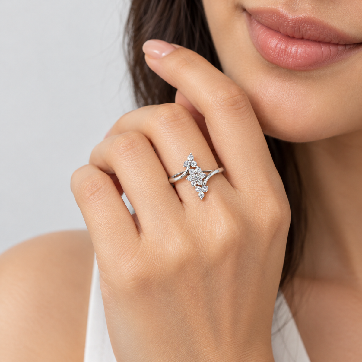 A close-up lifestyle shot of a woman's hand wearing a silver-toned ring with a delicate floral or leaf-like cluster of small clear stones, set against a soft light gray background.