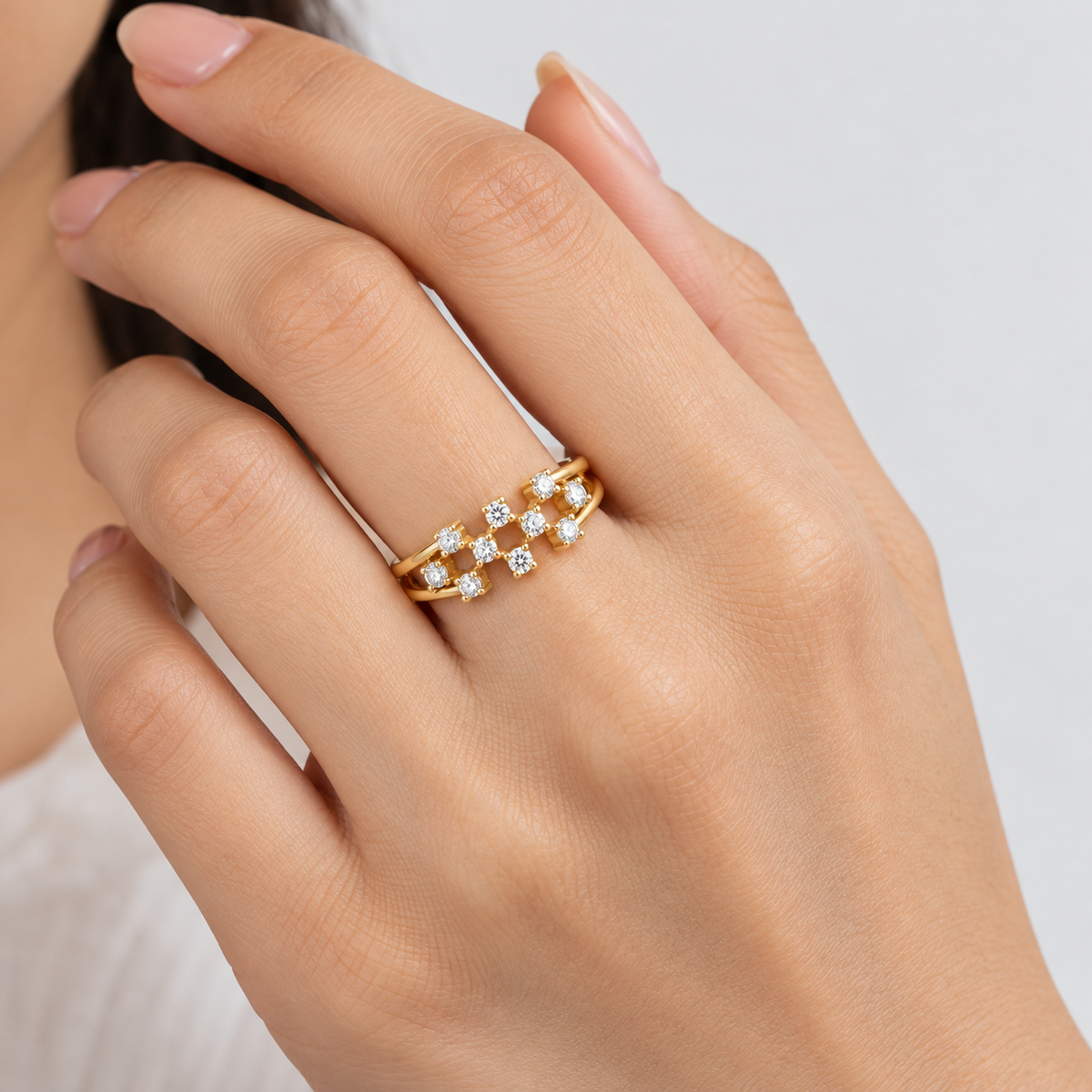 A close-up of a hand wearing a delicate gold ring set with small clear stones, photographed against a soft light gray background.