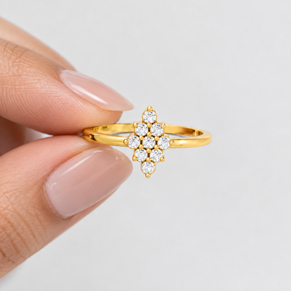 A gold ring with a floral cluster of clear gemstones held between fingers against a light gray background.