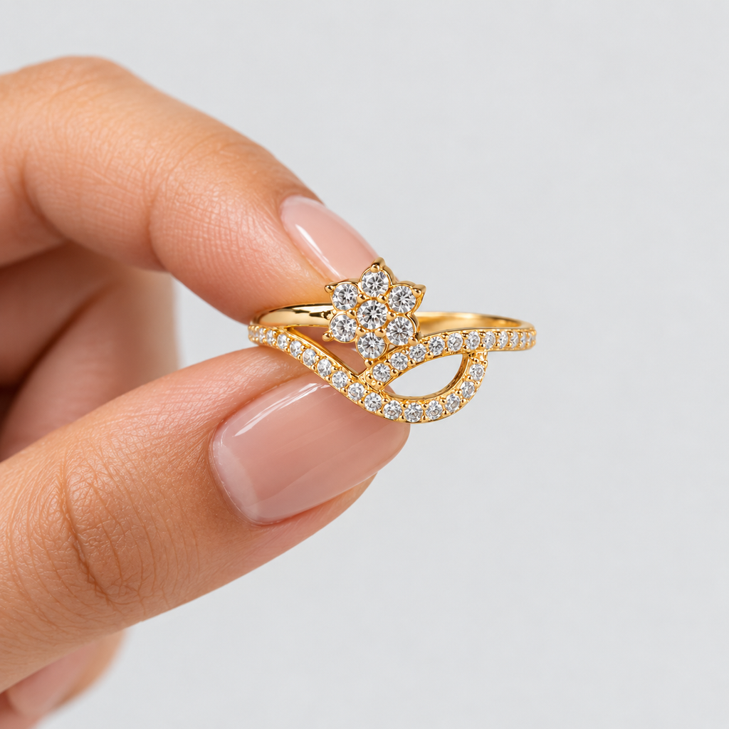 A gold ring with a flower-shaped cluster of clear stones and an intertwined curved band paved with small clear stones, shown being held between fingers against a light gray background.