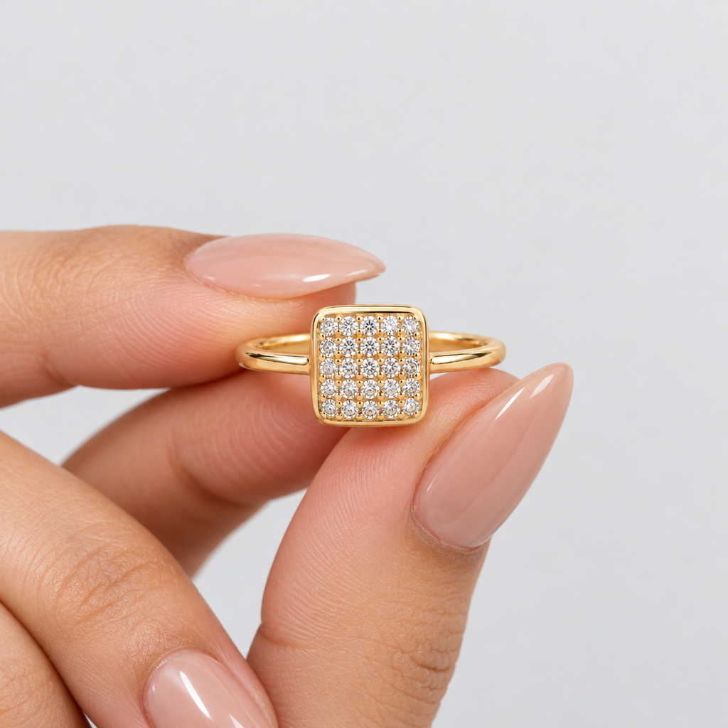 A gold square ring with a pavé diamond center, held between fingers against a light gray background.