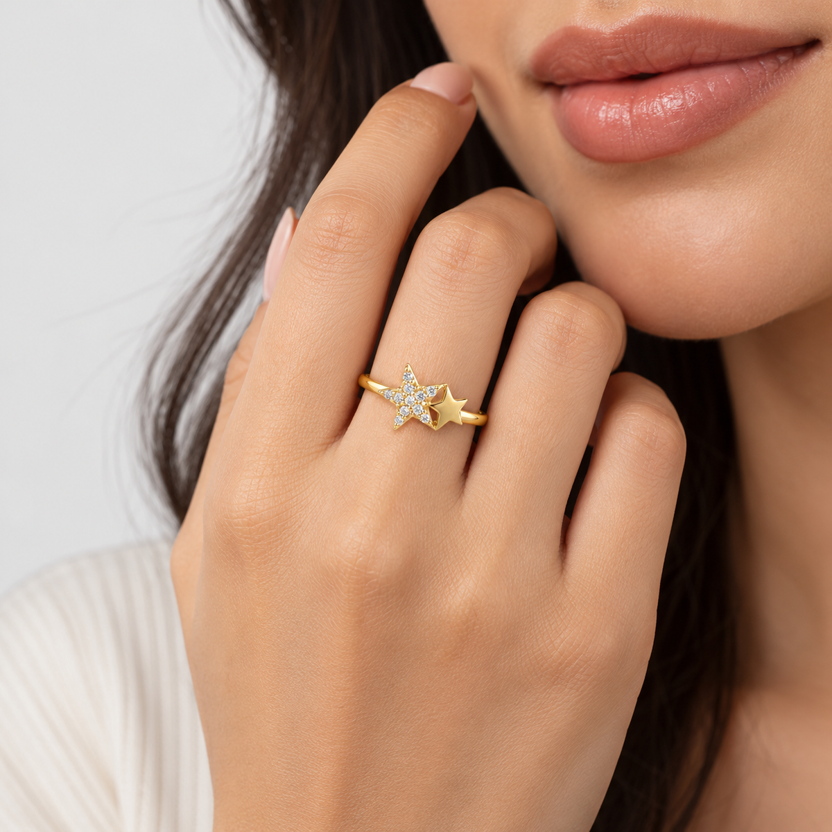 A close-up lifestyle shot of a model wearing a gold ring with a star design set with small clear stones. The model's hand, lips, and hair are visible against a clean, light gray background.