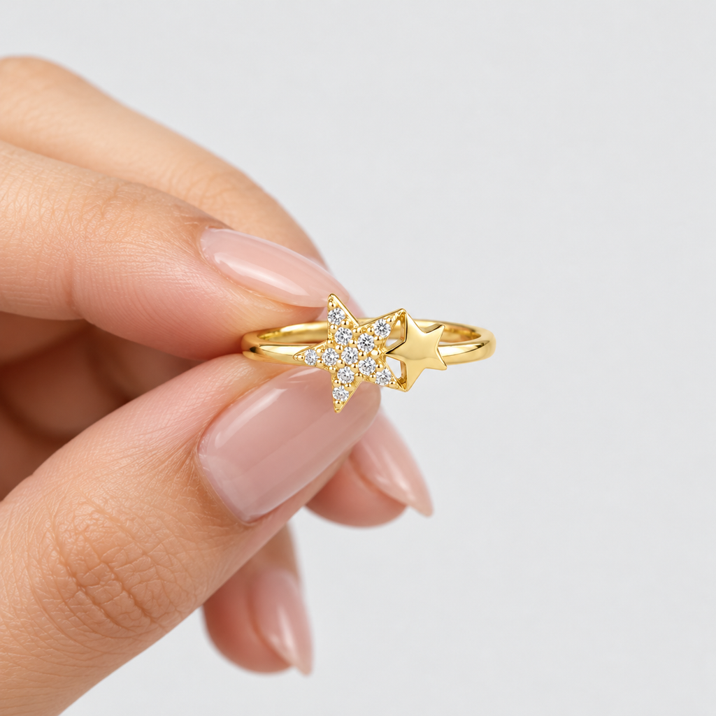 A gold ring featuring two star designs, one adorned with small clear crystals, held between fingers against a light gray background.