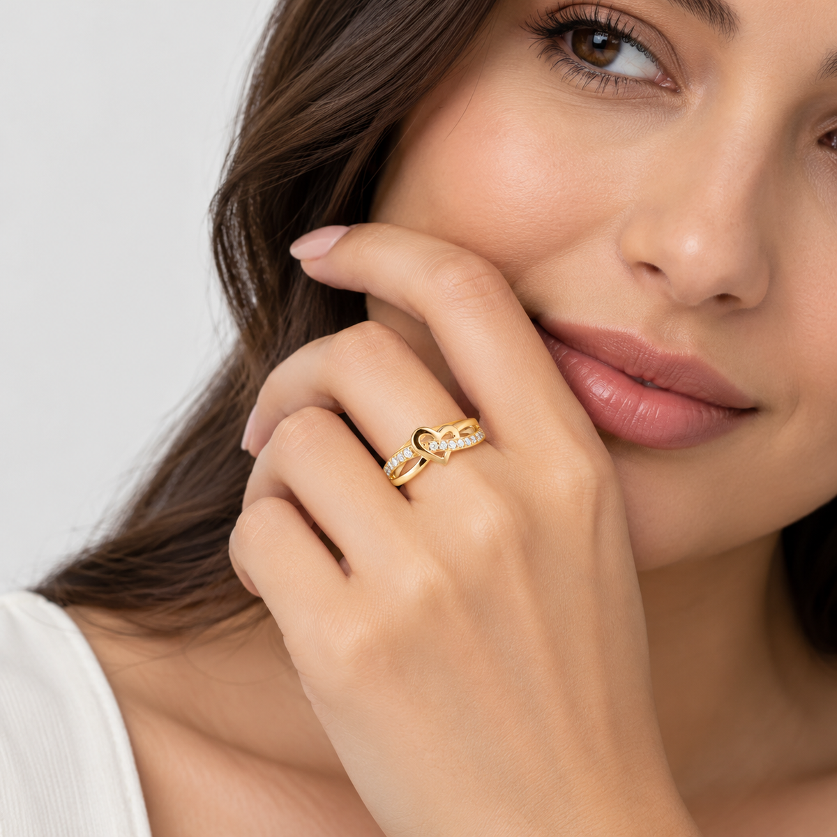 A close-up lifestyle shot of a woman wearing a delicate gold ring with a small heart-shaped design and tiny clear stones, resting her hand near her face against a soft light gray background.