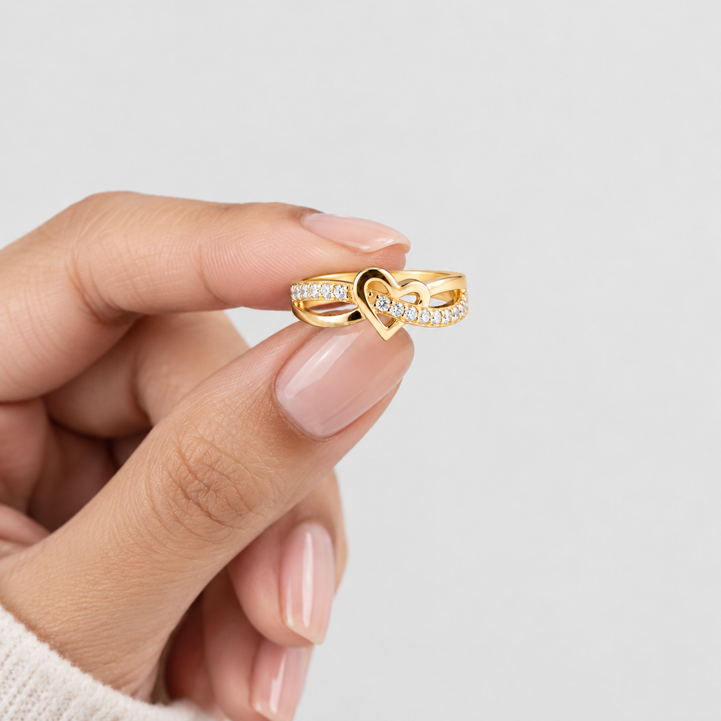 A gold ring with a heart-shaped cutout and pavé-set clear stones, held between fingers against a light gray background.