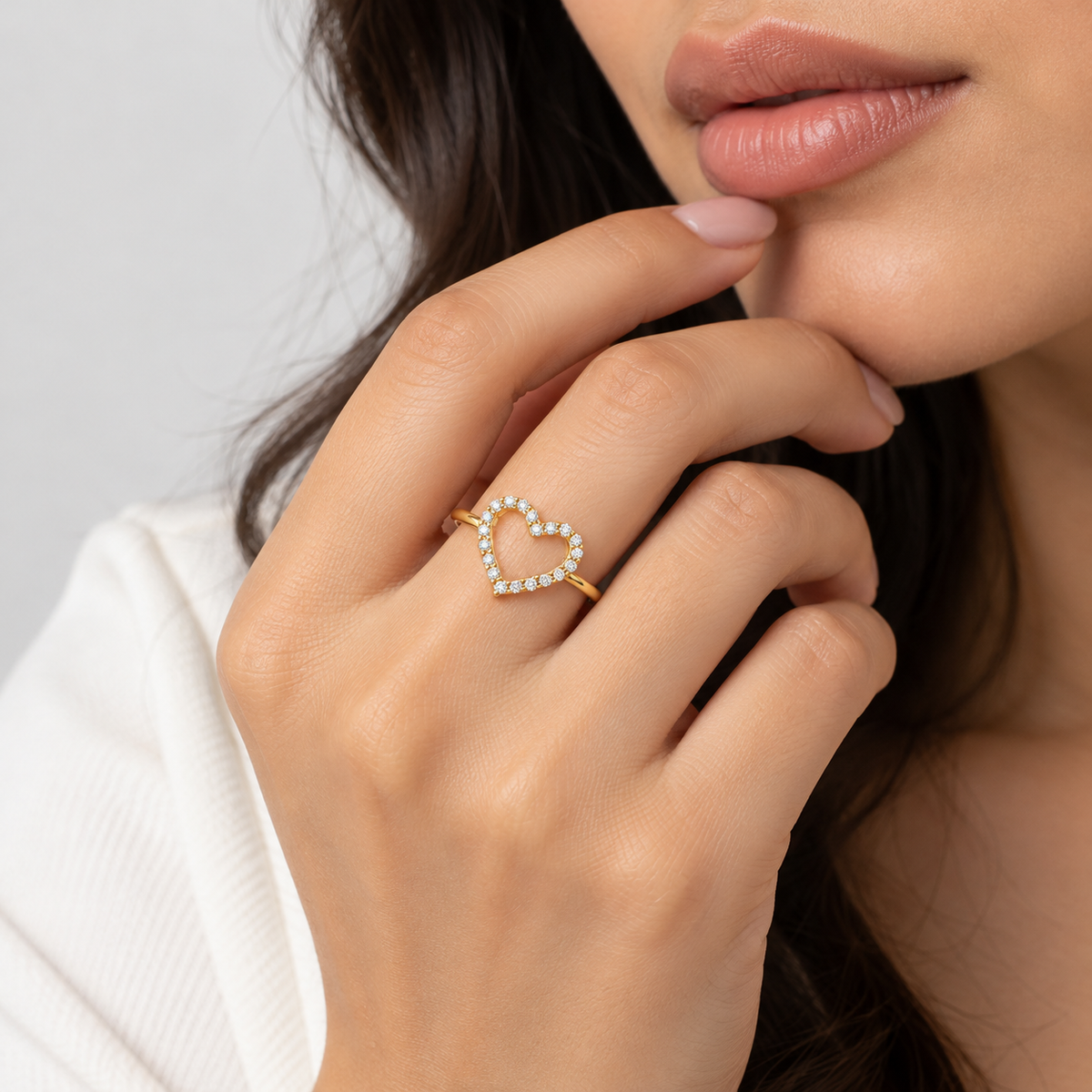 A close-up lifestyle shot of a woman wearing a delicate gold ring with a heart-shaped outline paved with small clear stones on her finger.