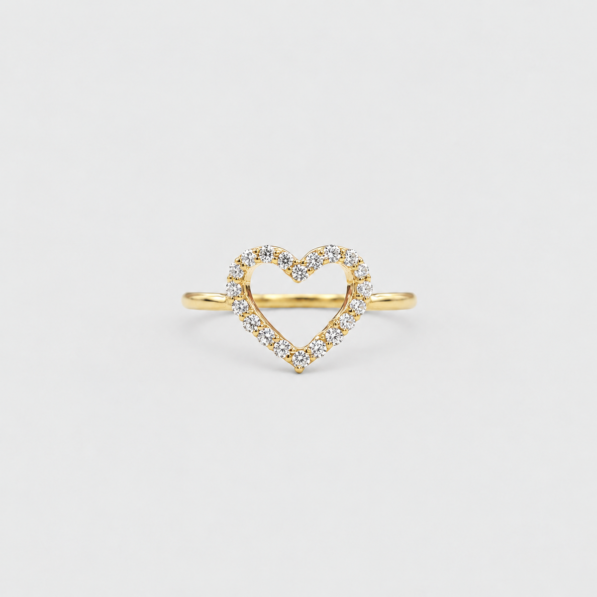 A delicate yellow gold ring featuring an open heart-shaped center paved with small clear stones, set against a solid light gray background.
