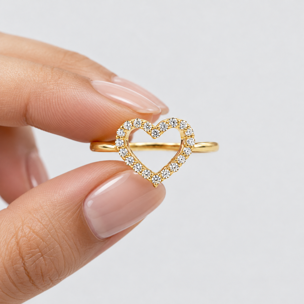 A gold heart-shaped ring with small clear stones, held between fingers against a light gray background.