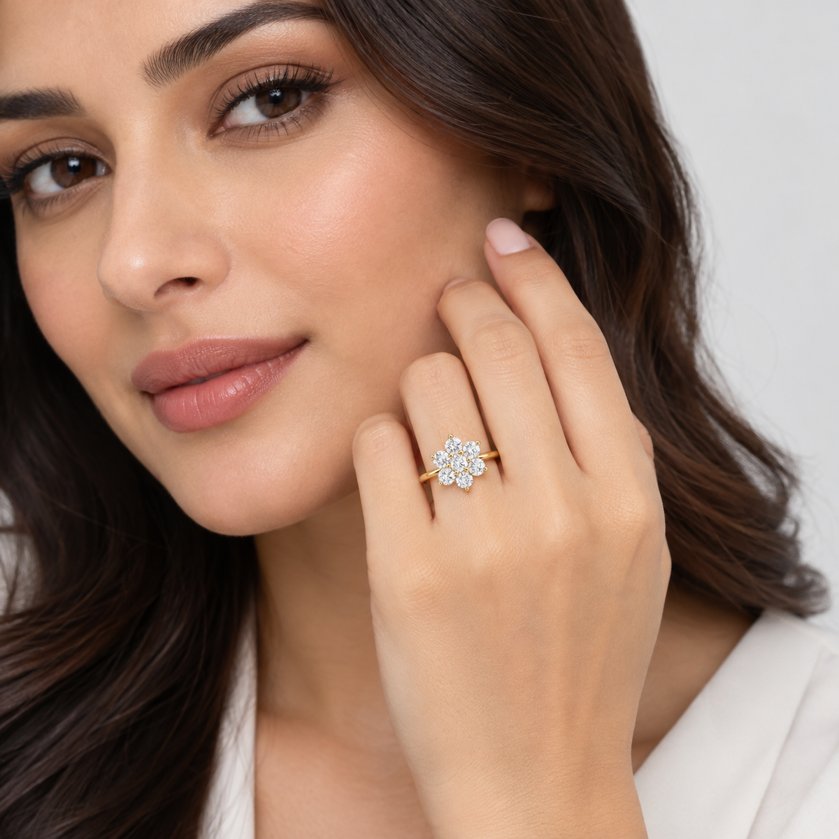 A close-up lifestyle shot of a woman wearing a gold ring with a flower-shaped cluster of clear gemstones on her finger, set against a clean, light gray background.