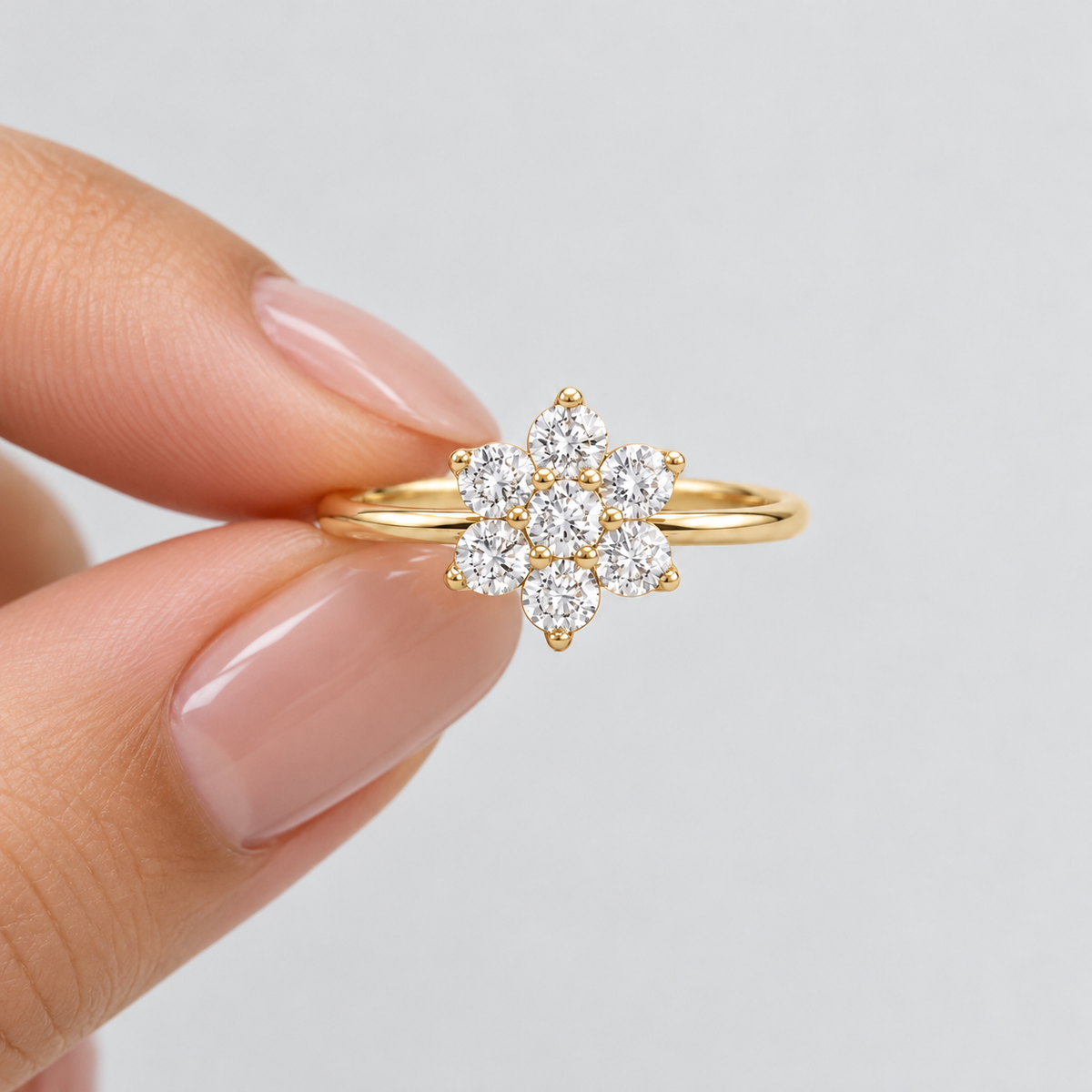 A gold ring with a flower-shaped cluster of clear gemstones, held between two fingers against a light gray background.