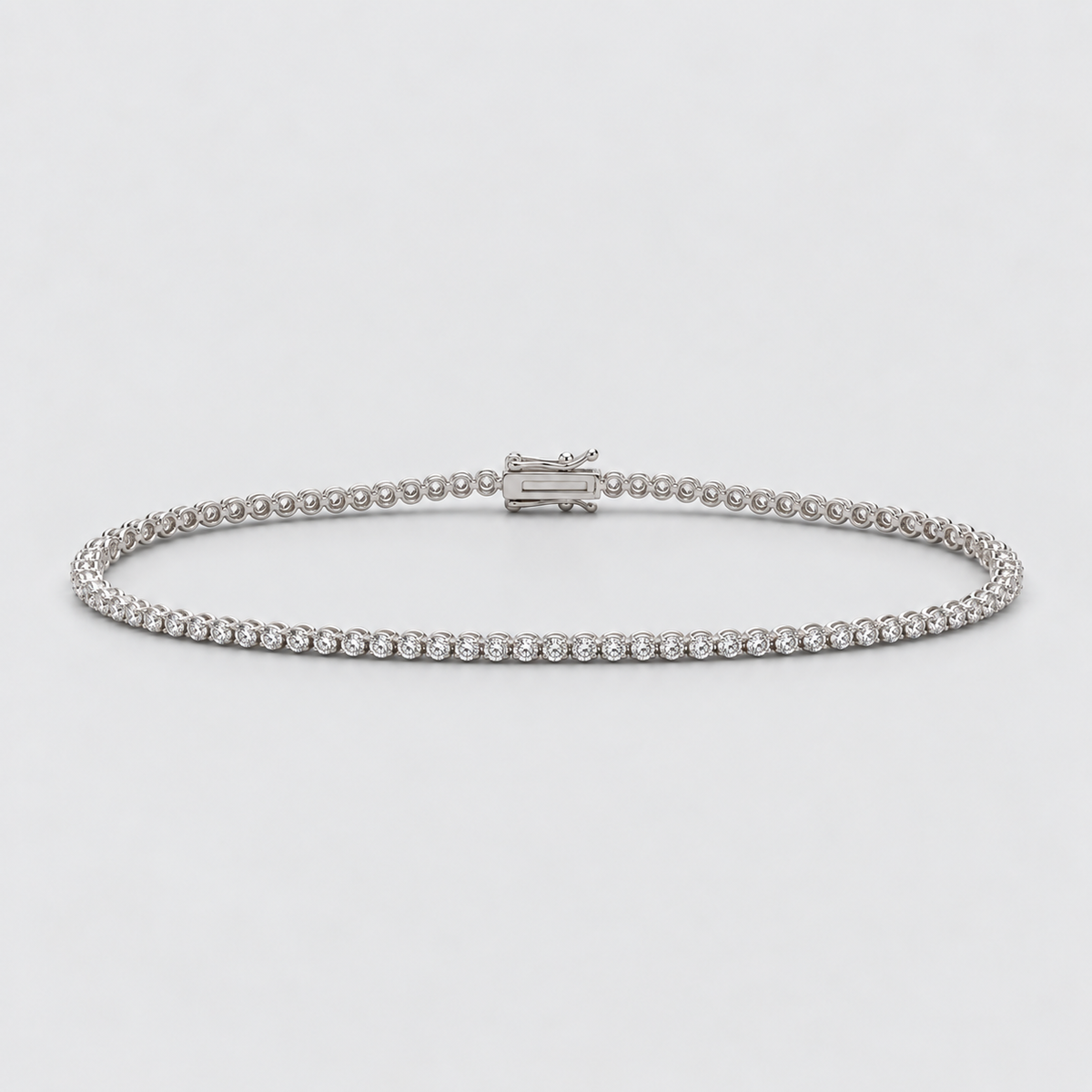 A silver tennis bracelet with a row of small clear stones and a rectangular clasp, photographed on a light gray background.