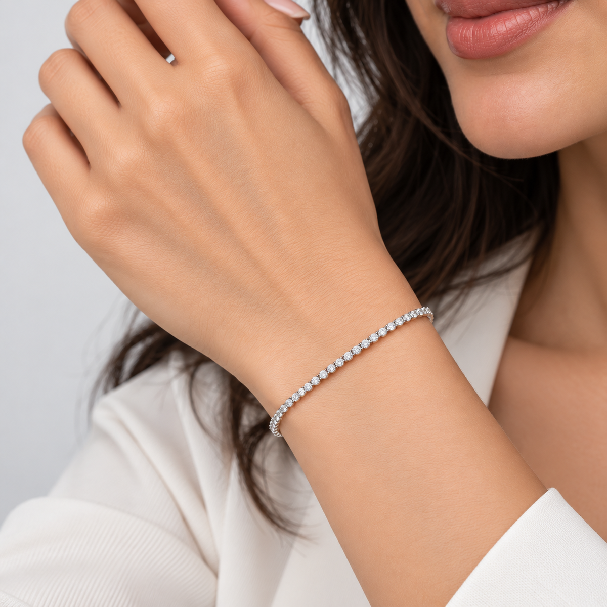 A close-up lifestyle shot of a woman wearing a delicate silver tennis bracelet on her wrist, with her hand resting near her face. She is wearing a white top, and a small silver ring is visible on her finger.