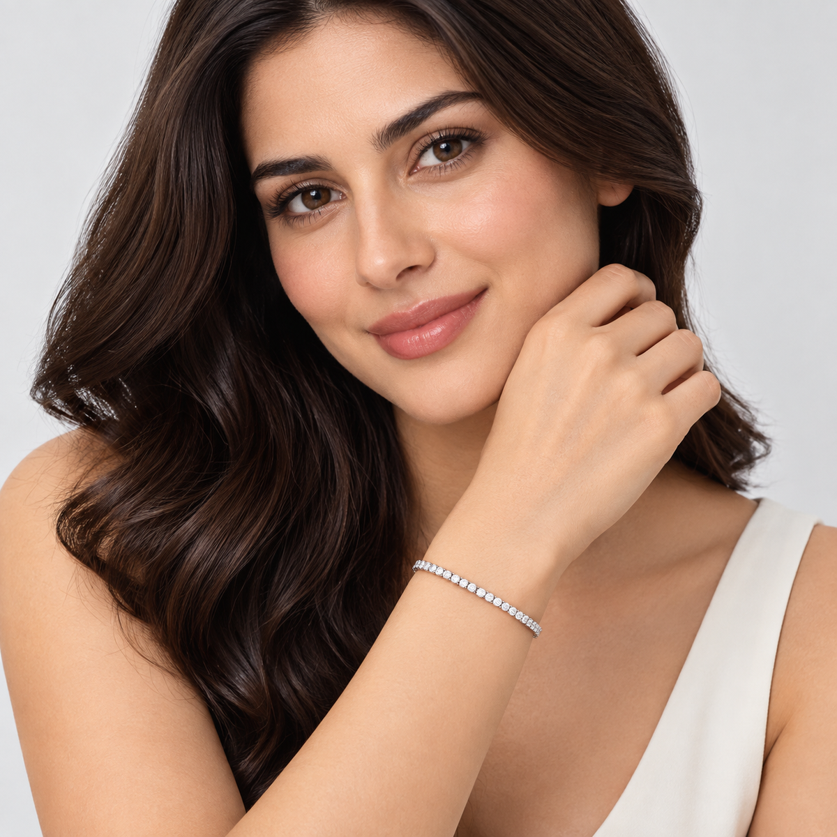 A woman with long dark brown hair models a delicate silver tennis bracelet against a clean, light gray background.