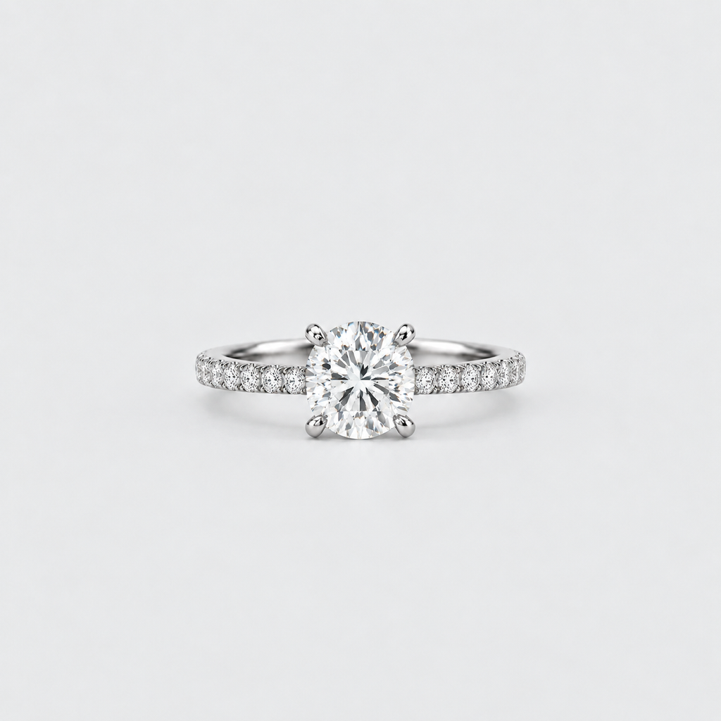 A classic solitaire engagement ring featuring a round brilliant-cut diamond set in a four-prong silver band with pavé diamonds on the shoulders, photographed on a clean light gray background.