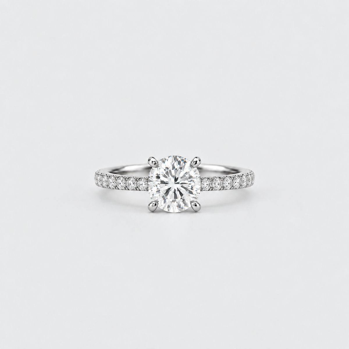A classic solitaire engagement ring featuring a round brilliant-cut diamond set in a four-prong silver band with pavé diamonds on the shoulders, photographed on a clean light gray background.