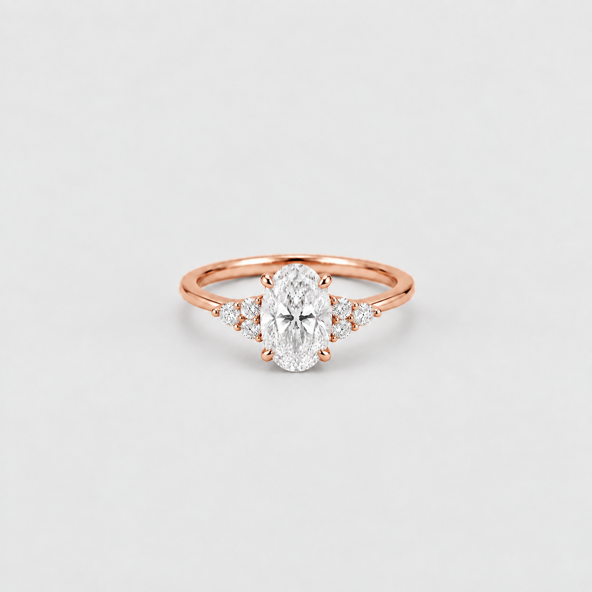 A rose gold engagement ring featuring an oval-cut center stone and small side stones, set on a plain light gray background.
