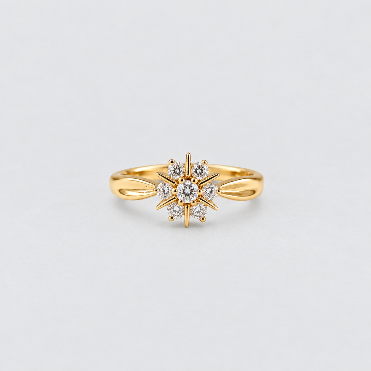 A gold ring featuring a starburst design with a central round clear gemstone surrounded by smaller clear stones, set on a plain light gray background.