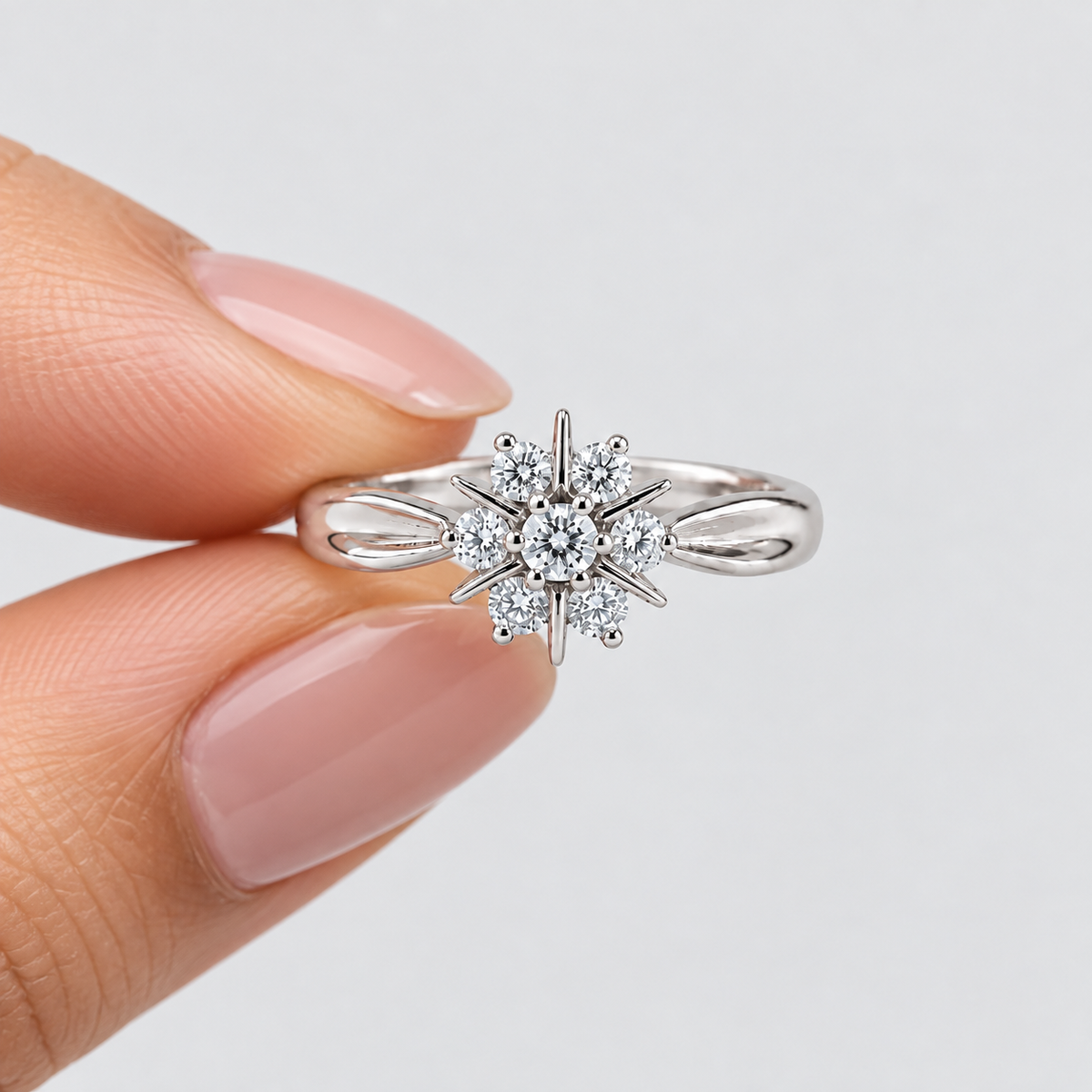 A silver-toned ring with a starburst cluster of clear gemstones, held between two fingers against a light gray background.