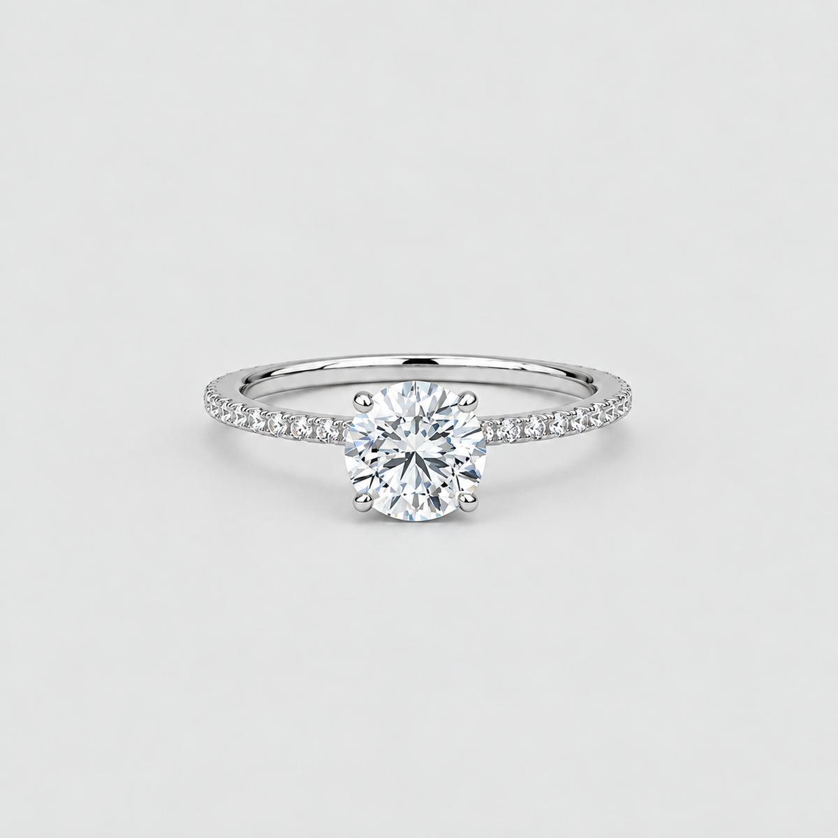 A classic solitaire engagement ring featuring a round brilliant-cut diamond set in a silver-colored metal band with a pavé band, photographed on a clean light gray background.