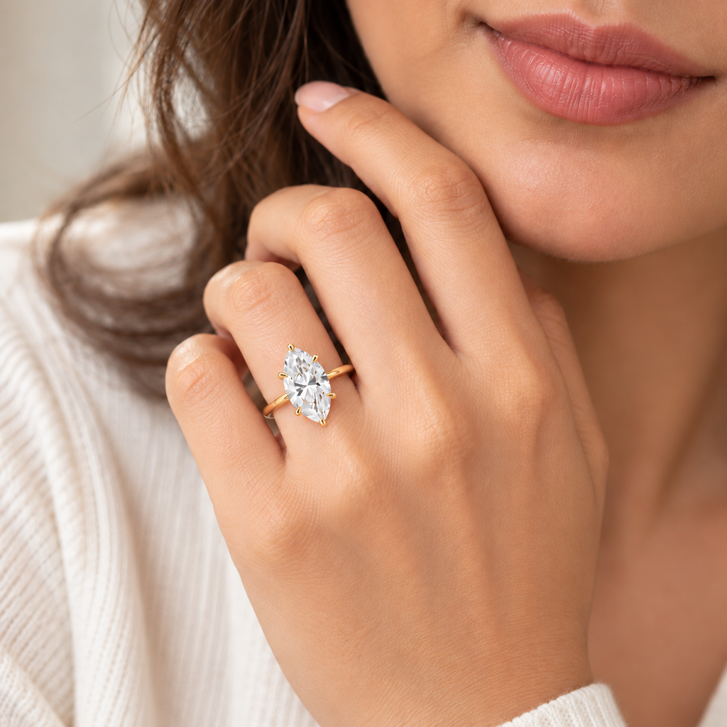 A close-up lifestyle shot of a woman wearing a gold ring with a pear-cut diamond on her finger. The ring is shown on her hand, which is resting near her face, against a soft, light-colored background.