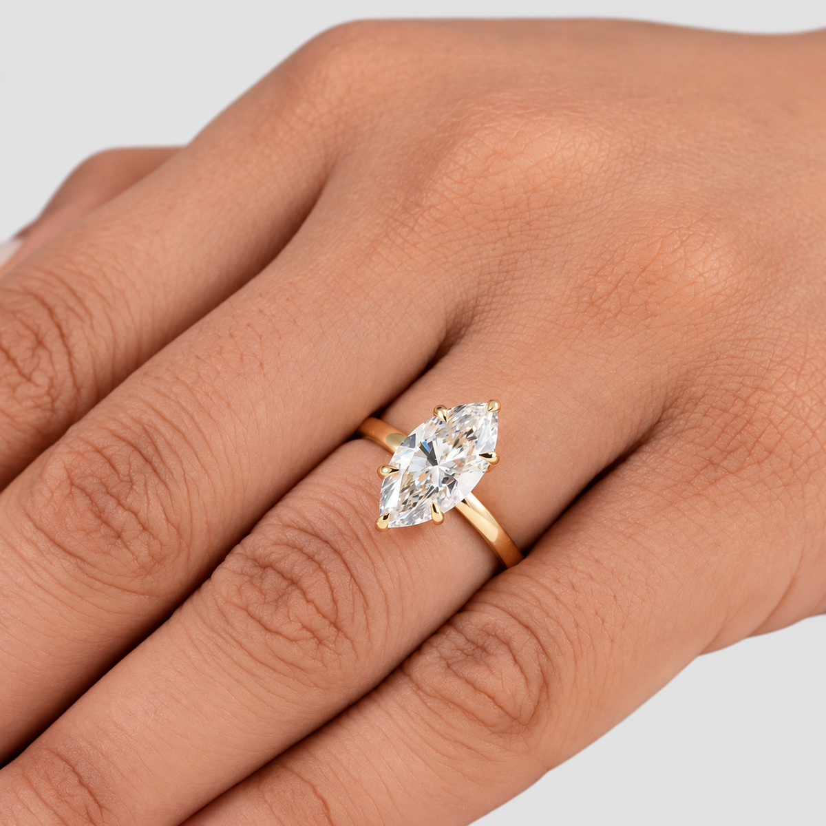 A close-up of a hand wearing a yellow gold ring with a marquise-cut clear gemstone, set against a plain light gray background.