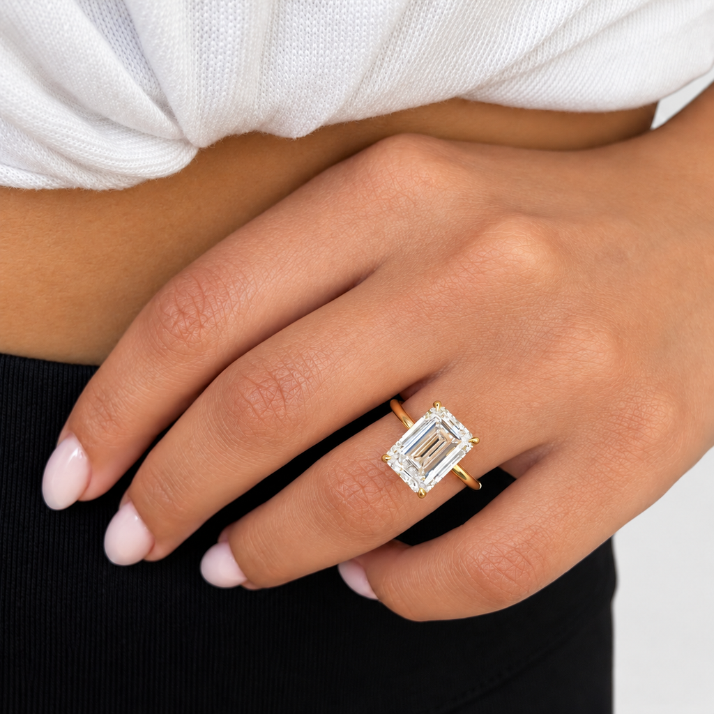 A close-up of a person's hand wearing a yellow gold ring with an emerald-cut clear center stone and smaller accent stones, set against a white top and black skirt background.