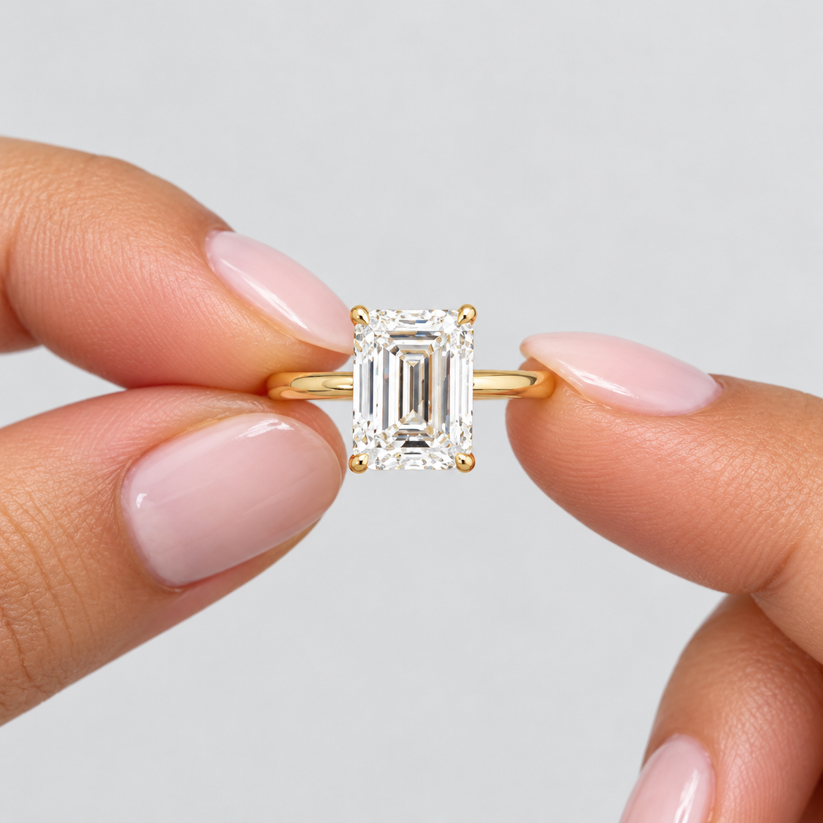 A close-up of a gold ring with an emerald-cut diamond held between two fingers against a light gray background.