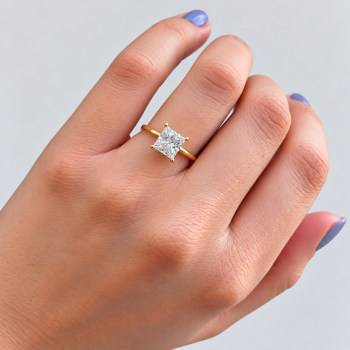 A close-up of a hand wearing a yellow gold ring with a princess-cut clear gemstone, set against a plain light gray background.