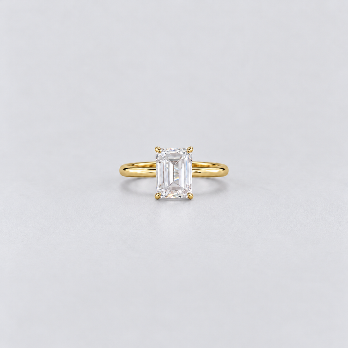 A yellow gold solitaire ring featuring an emerald-cut clear gemstone in a four-prong setting, centered on a light gray background.