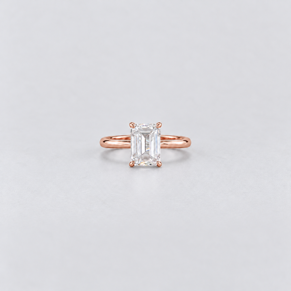 A rose gold engagement ring featuring an emerald-cut clear gemstone in a four-prong setting, centered on a light gray background.