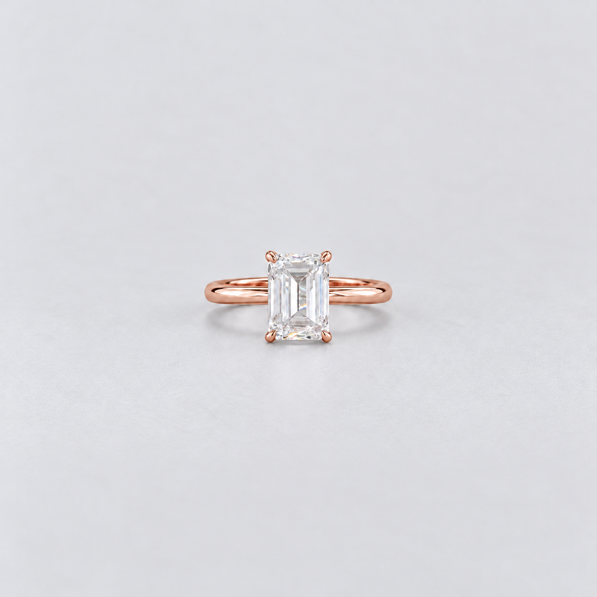 A rose gold engagement ring featuring an emerald-cut clear gemstone in a four-prong setting, centered on a light gray background.