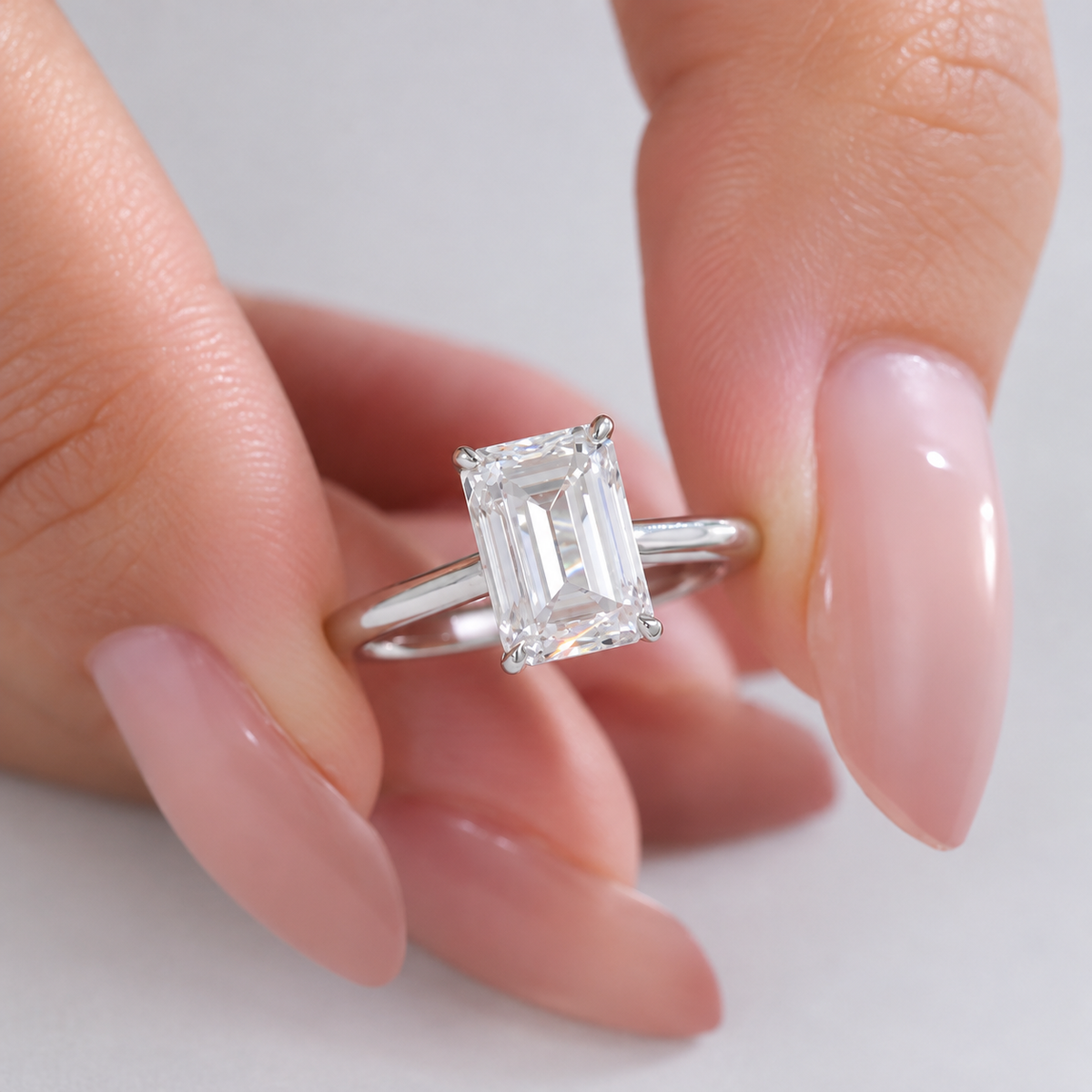 An emerald-cut diamond engagement ring on a silver band, held between two manicured fingers against a clean white background.