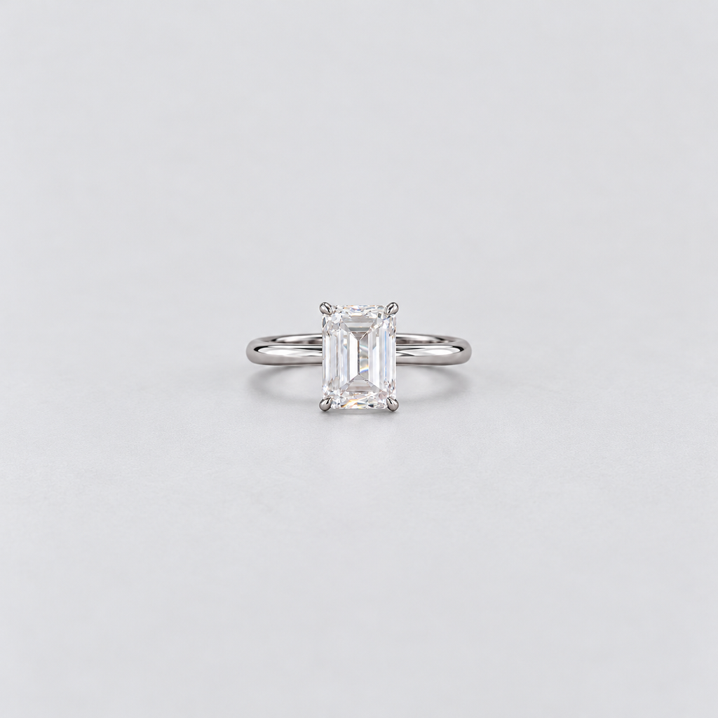 An emerald-cut diamond solitaire ring in a silver-colored metal band, centered on a light gray background.