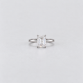 An emerald-cut diamond solitaire ring in a silver-colored metal band, centered on a light gray background.