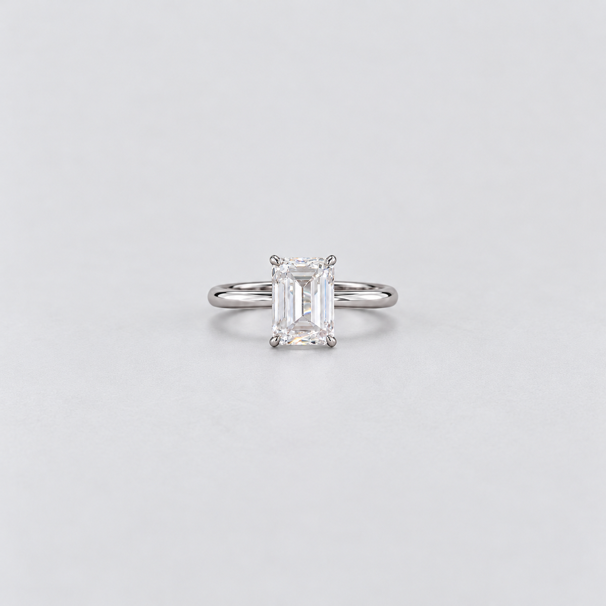 An emerald-cut diamond solitaire ring in a silver-colored metal band, centered on a light gray background.