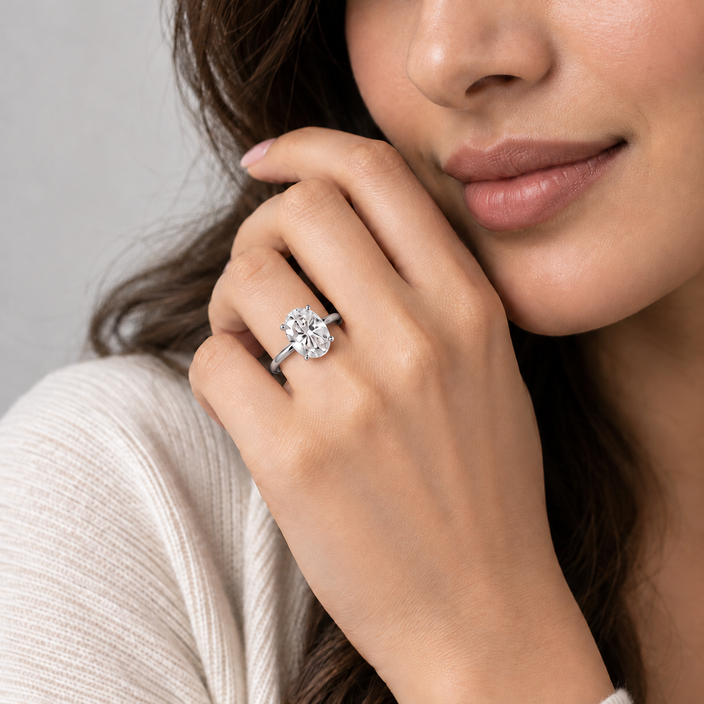 A close-up lifestyle shot of a woman wearing a classic solitaire engagement ring with a round brilliant-cut diamond on a silver band, set against a soft gray background.