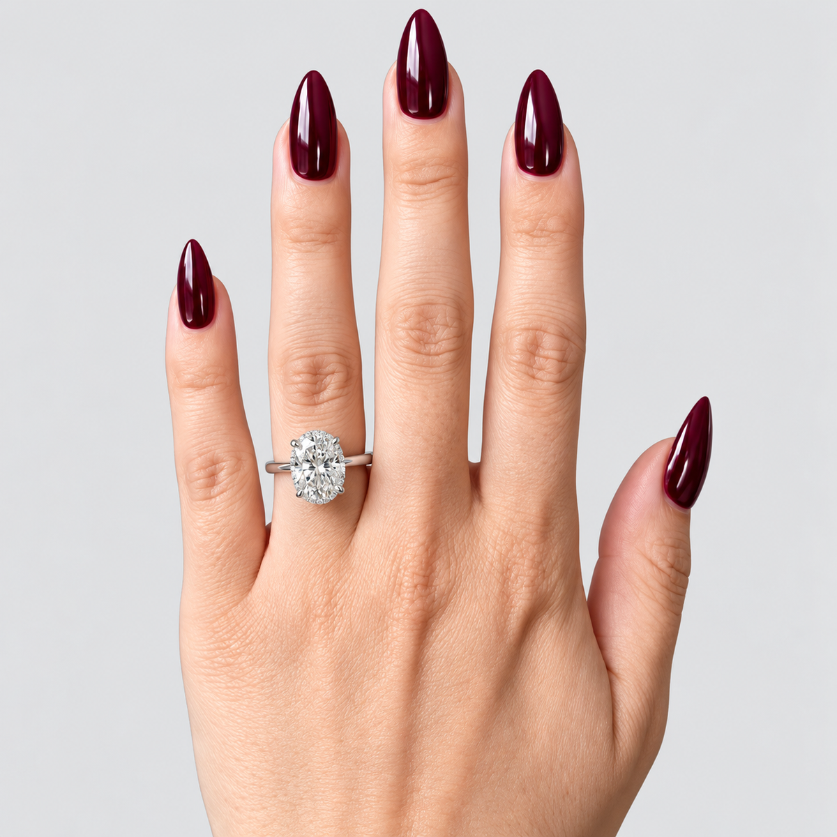 A close-up of a hand with long, glossy dark red manicured nails wearing a silver ring with a large oval-cut clear gemstone, set against a solid light gray background.