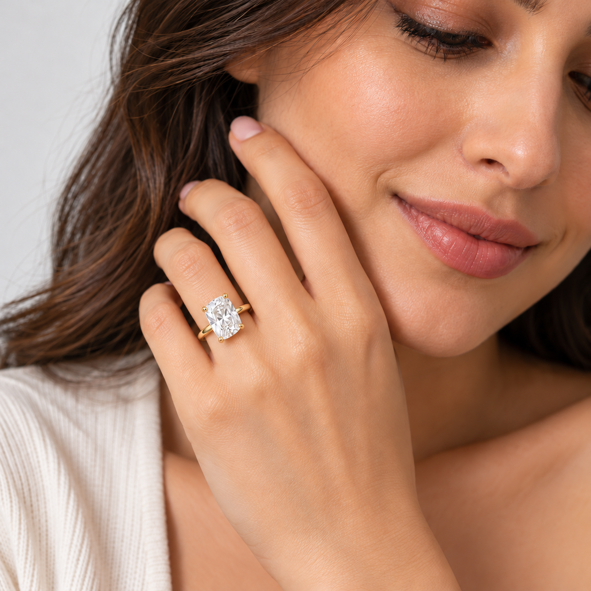 A close-up lifestyle shot of a woman wearing a yellow gold ring with a large oval-cut clear gemstone on her finger, with her hand resting against her cheek.