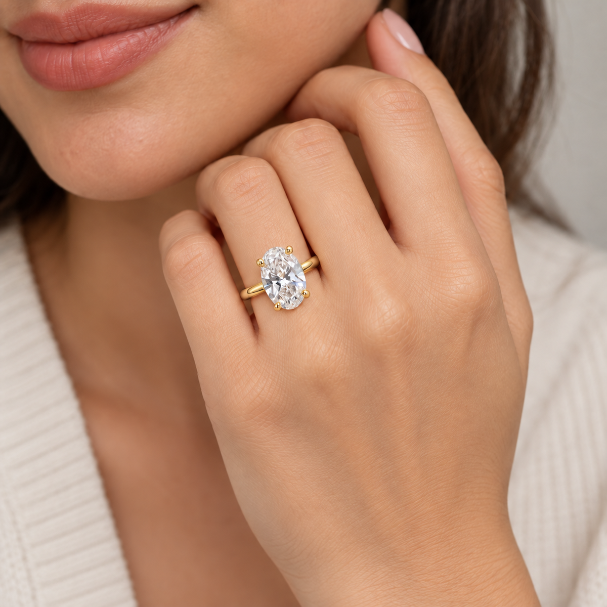 A close-up lifestyle shot of a woman wearing a yellow gold ring with an oval-cut clear gemstone on her finger, set against a soft neutral background.
