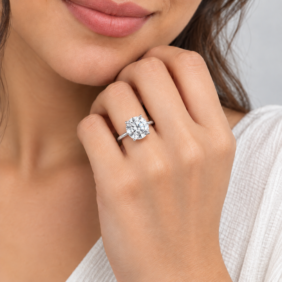 A close-up lifestyle shot of a woman wearing a silver-toned ring with a large cushion-cut diamond. The ring is shown on her finger, with her hand resting near her chin, wearing a white textured top against a neutral light gray background.