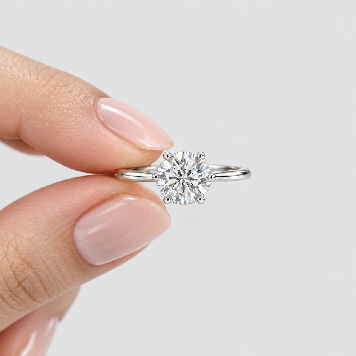 A close-up of a hand with pale pink manicured nails holding a classic solitaire engagement ring. The ring features a round brilliant-cut diamond set in a polished silver-colored band, photographed against a clean, light gray background.