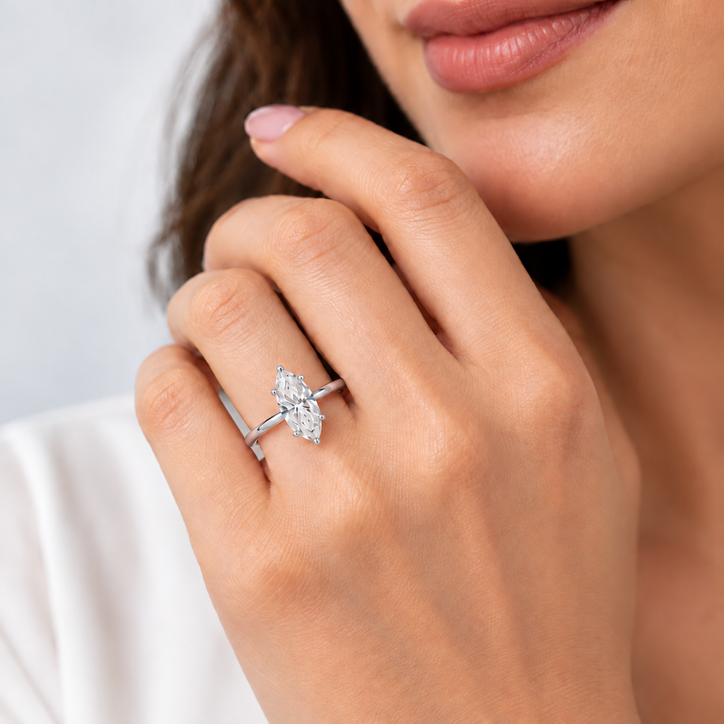 A close-up lifestyle shot of a woman's hand wearing a silver-toned diamond ring with a marquise-cut diamond cluster, set against a soft, light gray background.
