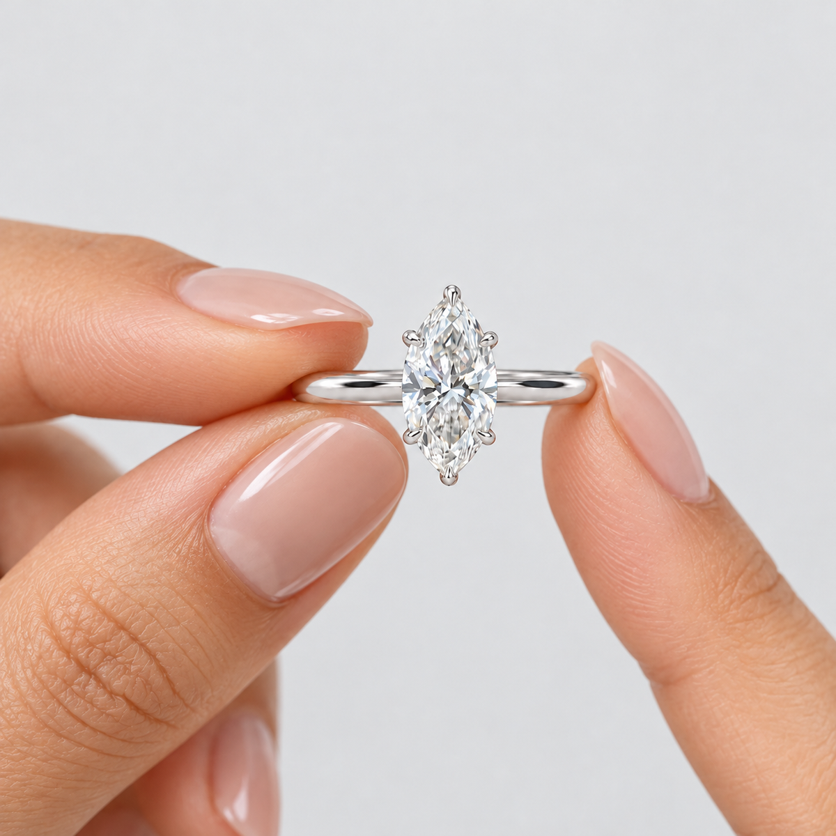 A close-up of a hand with manicured nails holding a silver engagement ring with a marquise-cut diamond in a prong setting against a light gray background.