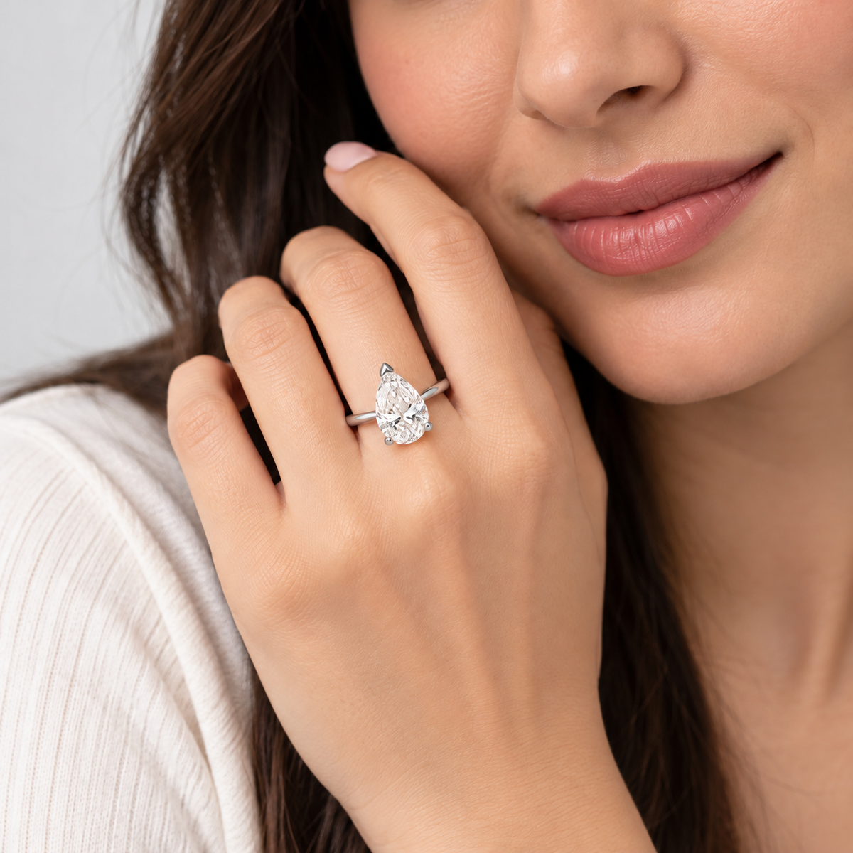 A close-up lifestyle shot of a woman wearing a pear-cut diamond ring on a silver band, resting her hand near her face.