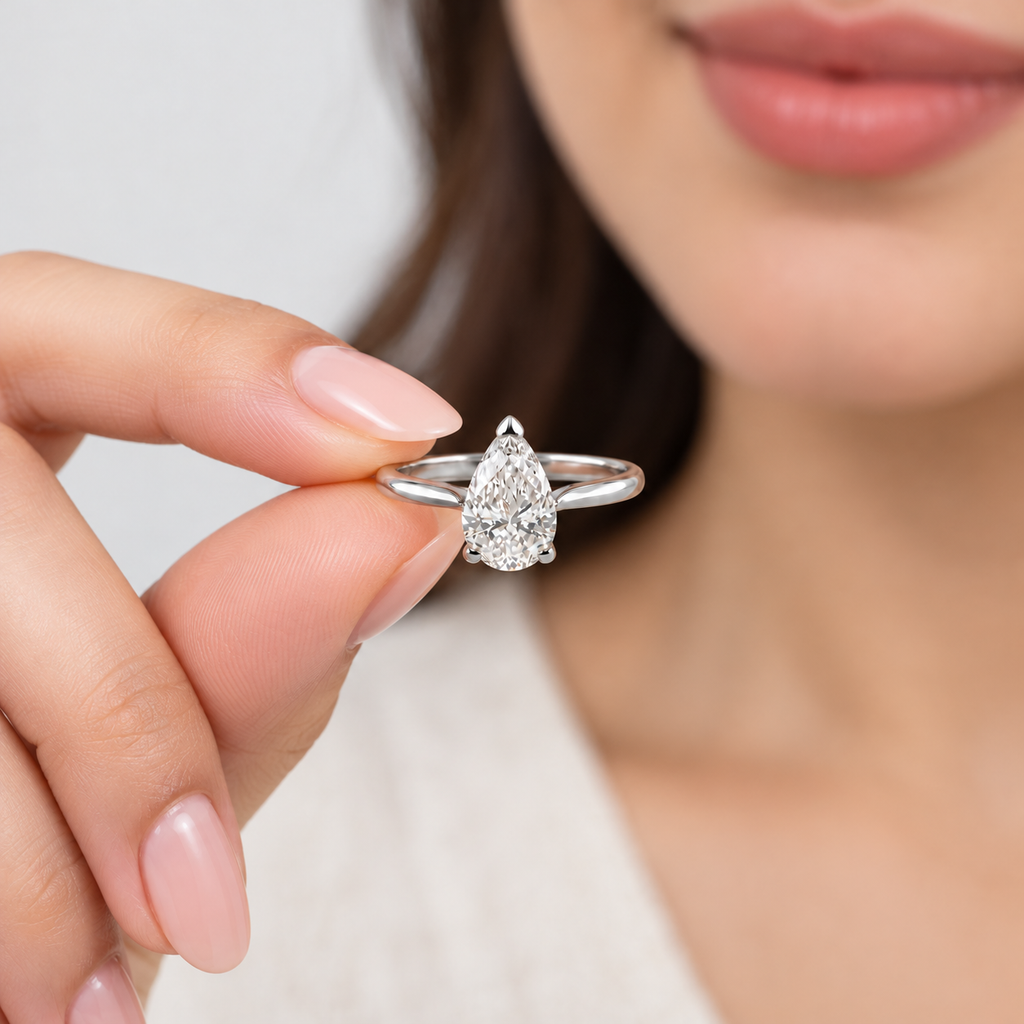 A close-up lifestyle shot of a woman holding a silver ring with a pear-cut clear gemstone between her fingers. The background is a soft, out-of-focus light gray.